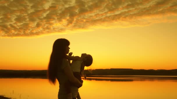 Mother plays with her baby by water. happy family concept. silhouette of mother and healthy baby circling. mom tosses her happy little daughter up in air at beach while having fun at sunset. — Stock Video