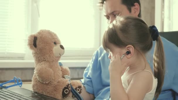 Niña en la oficina de un pediatra, juega y trata juguete osito de peluche y lo escucha con estetoscopio. pediatra masculino jugando con un niño en el consultorio. bebé niño se sienta en el regazo del pediatra — Vídeo de stock