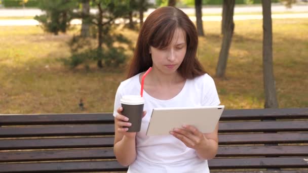 Chica en un parque de verano, la cafetería utiliza tabletas y bebidas delicioso café aromático. mujer sentada en el banco charlando, tomando café y descansando. mujer joven en la calle trabaja con un gadget y bebe té — Vídeo de stock