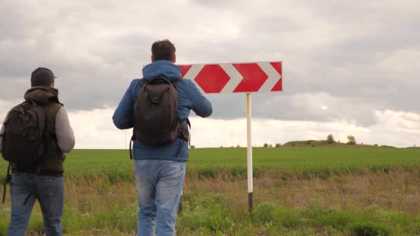 Reisende mit Rucksack kommen zum Verkehrszeichen an der Kreuzung und wählen ihre Route. Teamwork der Touristen. Wander- und Erlebniskonzept. Männer sind verloren, auf der Suche nach der richtigen Reiserichtung. — Stockvideo