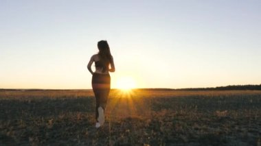 Sağlıklı, güzel bir kız spor yapıyor, kırsalda güneş altında koşuyor. Koşucu kız sahada temiz hava soluyor. Özgür genç bir kadın gün batımında yaz parkında koşar. Güneşin peşinden koşmak. eğitim koşusu.