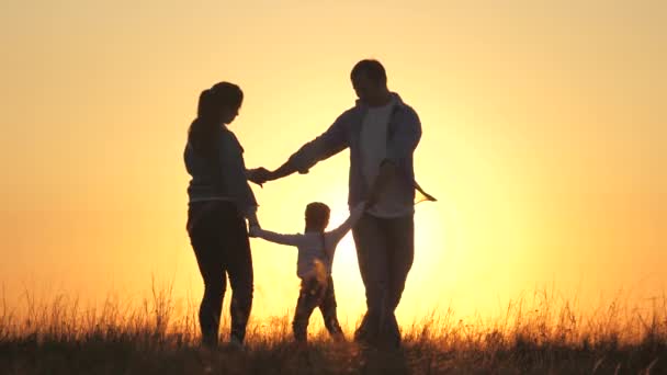 Mamá joven, papá con una hija sana bailar en un círculo bajo el sol caliente, divertirse en el campo. familia feliz, tomados de la mano, juega en el parque en la hierba, al atardecer. Concepto de familia e infancia. — Vídeo de stock