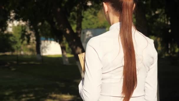 Een student loopt door de straat met leerboeken in haar handen in het zomerpark. schoolmeisje in de stad. meisje tiener haast naar school met boeken. — Stockvideo