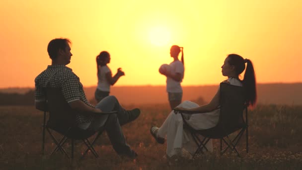 Feliz familia viajeros mamá papá y los niños juegan al atardecer en el parque. mamá y papá están sentados en sillas turísticas en el campo los niños están jugando pelota en el sol caliente. concepto de una familia feliz y la infancia. — Vídeo de stock