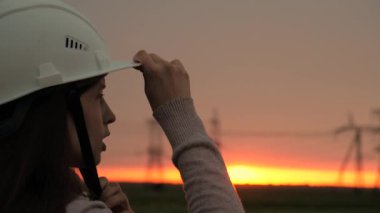 Beyaz kasklı bir kadın güç mühendisi tabletteki elektrik sensörlerinden elde edilen verileri kullanarak güç hattını kontrol ediyor. Gün batımında yüksek voltajlı elektrik hatları. Dağıtım ve elektrik tedariği. temiz enerji