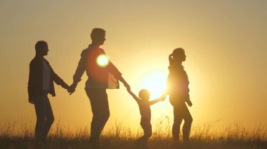 Mutlu yedinin silueti, el ele yürüyor, gün batımında tarlada yürüyor. Çocuklar anne ve babalarının elini tutuyor. Mutlu küçük çocuk ve ebeveynler güzel güneş ışınlarında yürürler, tatile çıkarlar. a