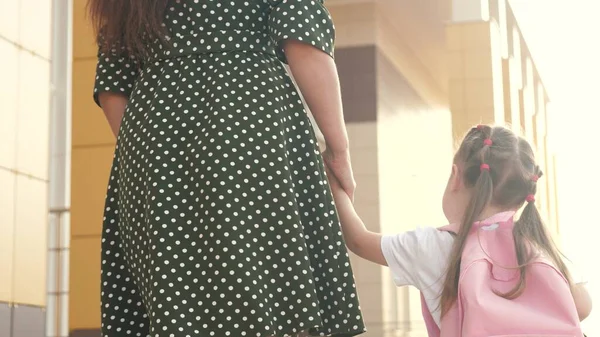 Kleine Tochter Schulmädchen geht mit einem Rucksack und hält ihre Mutter Hand. Vorschulpädagogisches Konzept. Glückliche Familienmutter und Kind gehen Händchen haltend zur Schule. — Stockfoto