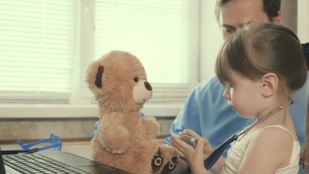 子供は聴診器で医者の言うことを聞く。小児科医の膝の上に座っている患者の少女。専門的な男性小児科医は、オフィスで小さな子供と遊ぶ介護. — ストック動画