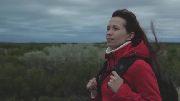 Una chica viajera despreocupada mira a la distancia, el viento agita su cabello. amor por la aventura y los viajes. Mujer libre viaja con mochila, mira desde lo alto de la colina en el bosque, disfrutando del paisaje. — Vídeos de Stock