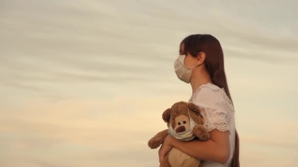 Niña lleva en su juguete de mano osito de peluche con máscara médica en el parque de verano. una mujer joven lleva el juguete favorito de su hijo. juguete osito de peluche en máscara médica. Covid-19 pandemic coronavirus. concepto de salud — Vídeo de stock