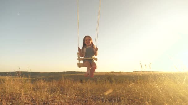 Zdravé šťastné dítě jezdí na provaze na větvi v parku. veselé děvčátko se směje, raduje se a mává rukama za letu. Máma třese dcerou na houpačce pod stromem na slunci. Dětství a — Stock video