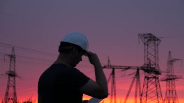 Beyaz kasklı erkek güç mühendisi güç hattını kontrol ediyor, elinde bir tablet tutuyor. Gün batımında yüksek voltajlı elektrik hatları. Dağıtım ve elektrik tedariği. temiz enerji