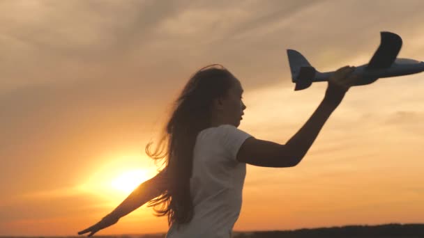 Gelukkig meisje rent met een speelgoedvliegtuigje op een veld bij zonsondergang. Het meisje wil piloot en astronaut worden. Langzame beweging. Kinderen spelen speelgoed vliegtuig. Tiener droomt van vliegen en piloot worden. — Stockvideo