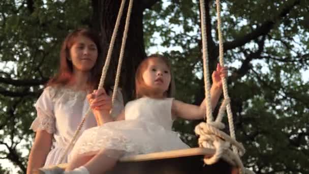 Mutter schüttelt ihre gesunde kleine Tochter auf einer Schaukel unter einem Baum in der Sonne. Mutter spielt mit Kind, sie schwingen am Seil auf einem Eichenzweig im Wald. Die freie Familie hat Spaß im Park. — Stockvideo