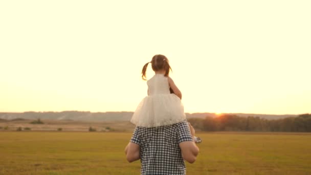 Far går med sin datter på skuldrene i solnedgangsstråler. Far bærer på skuldrene af sit elskede barn, i solstråler. barn med forældre går ved solnedgang. lykkelig familie hviler i parken. – Stock-video