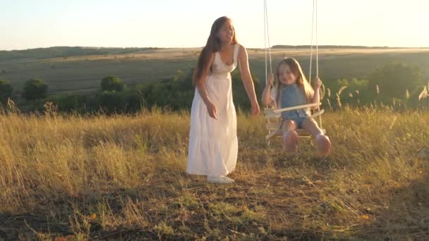 Mãe brinca com seu bebê, eles balançam em uma corda em um galho de árvore contra um céu azul. mamãe sacode sua filhinha saudável em um balanço sob uma árvore ao sol. família livre está se divertindo no parque. — Vídeo de Stock