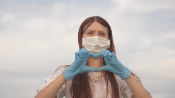 Meisje in een beschermend masker en medische handschoenen toont hart symbool met haar handen. Jonge vrouw houdt van dokters. pandemie, covid 19. vrouwelijke arts toont vingers van het hart en liefde voor patiënten. — Stockvideo