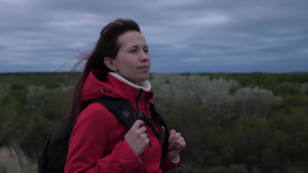 Una chica viajera despreocupada mira a la distancia, el viento agita su cabello. amor por la aventura y los viajes. Mujer libre viaja con mochila, mira desde lo alto de la colina en el bosque, disfrutando del paisaje. — Vídeos de Stock