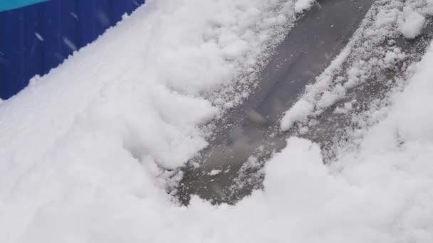 El conductor saca el coche de la nieve cerca de la casa. Raspando nieve y hielo del parabrisas de un coche. Una mujer limpia el coche de nieve con un cepillo. Las nevadas cubrieron el coche. — Vídeos de Stock