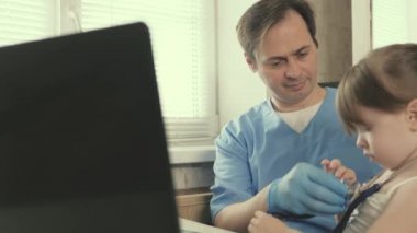 Bir çocuk doktoru tarafından küçük bir çocuğa bakılır. Doktor steteskopla çocuğun kalbini dinler. Özel klinikler. Ofiste küçük bir çocukla oynayan profesyonel bir erkek çocuk doktoru.