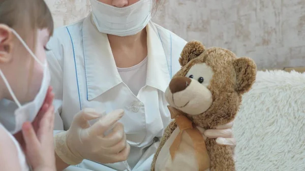 Menina bonito faz uma injeção para urso de brinquedo. bebê brinca com uma máscara médica. criança brinca no hospital. jogo finge ser médico, enfermeiro, veterinário, cura um paciente com uma vacina. perigo de coronavírus — Fotografia de Stock