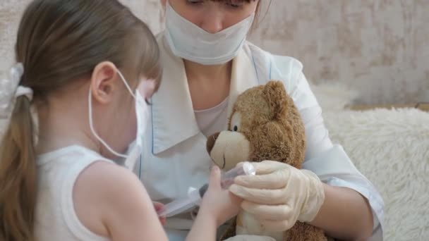 Linda chica hace una inyección para oso de juguete. bebé juega con una máscara médica. niño juega en el hospital. juego pretende ser médico, enfermera, veterinario, cura a un paciente con una vacuna. peligro de coronavirus — Vídeos de Stock