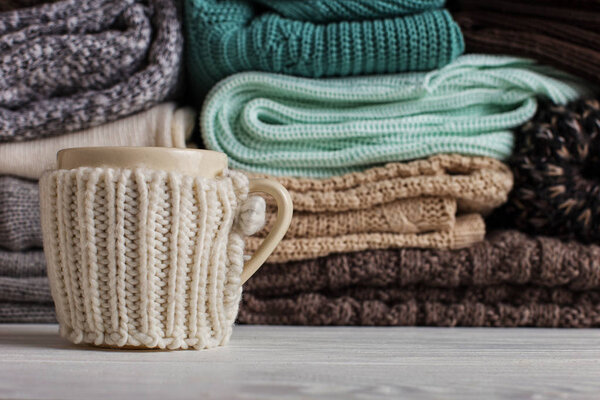 A stack of knitted clothes of different colors and textures, on the table next to a cup in a case.