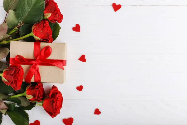 A bouquet of red roses, gift and hearts on white wooden table. Concept Women's Day or St. Valentine. Copy space.