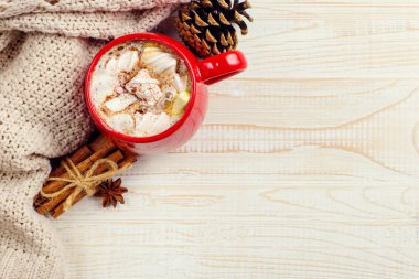 Kış kahvesi, kremalı kakao ve kırmızı seramik fincanda marşmelov. Tahta bir masanın üzerinde, kareli bir eşarbın yanında. Metin için yer, kopyalama alanı.