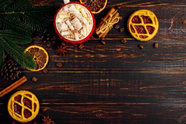 A cup of hot drink with whipped cream, marshmallows and powder, in a knitted cover and homemade cookies, cinnamon, Christmas decoration, lie on a wooden table. Copy space.