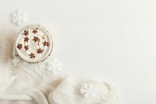 A cup of hot winter drink with whipped cream and a dusting powder with an asterisk, white snowflakes and knitted scarf on a wooden table. Christmas concept in bright colors. Copy space.