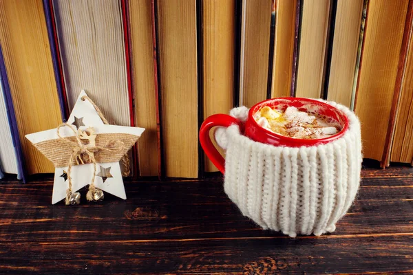 Winter coffee drink, cocoa with whipped cream and marshmallows in a red ceramic cup. Standing on the bookshelf, near the Christmas decoration star. Place for text, copy space.