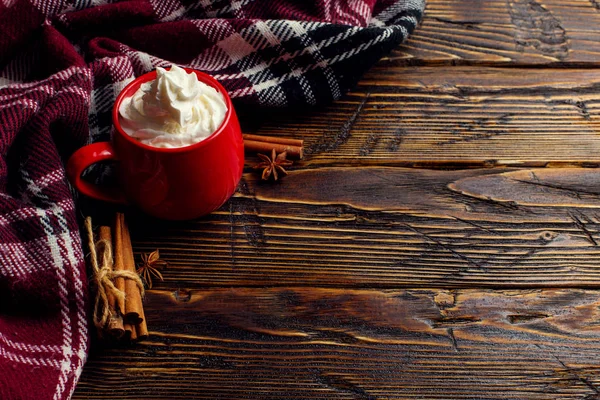 Winter coffee drink, cocoa with whipped cream and marshmallows in a red ceramic cup. Standing on a wooden table, next to a checkered scarf. Place for text, copy space.