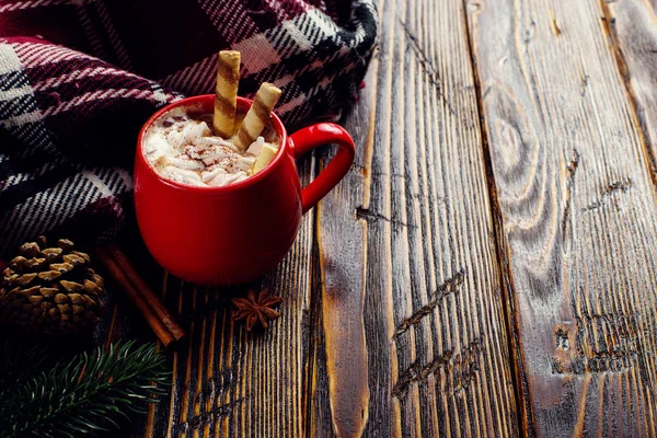 Winter coffee drink, cocoa with whipped cream and marshmallows in a red ceramic cup. Standing on a wooden table, next to a checkered scarf. Place for text, copy space.