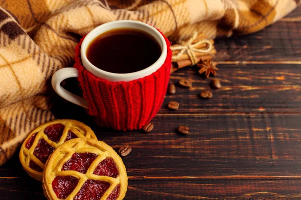 A cup of hot coffee, in a knitted cover and homemade cookies and spices, lie on a wooden table near the checkered plaid. Copy space.