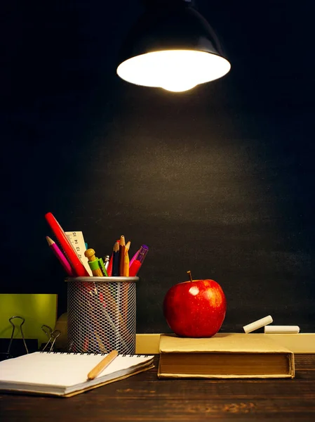 The teacher's desk or a worker, on which the writing materials lie, a book and an apple, in the evening under the lamp. Blank for text or background for a school theme. Copy space.