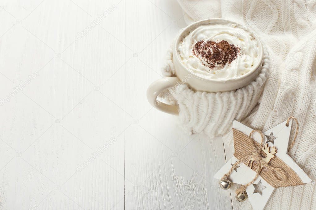 A cup of hot winter drink with whipped cream and a dusting powder with an asterisk, white snowflakes and knitted scarf on a wooden table. Christmas concept in bright colors. Copy space.