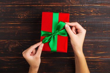 Dişi eller yeşil kurdeleli kırmızı bir hediye tutarlar. Ahşap bir masanın arka planında, üst görünüm.