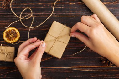 Women's hands making a gift of craft paper. Against the background of dried orange, cinnamon, pine cones, anise on a wooden table.