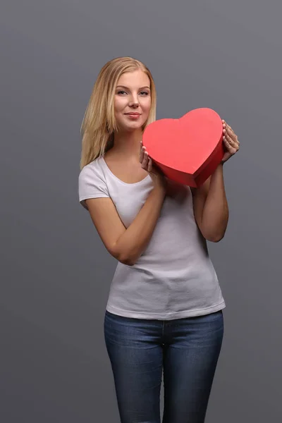 Young blonde girl with gift in hand, red in the shape of a heart. Emotions of joy and surprise on the face. Isolate on a gray background.