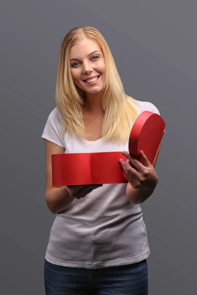 Young blonde girl with gift in hand, red in the shape of a heart. Emotions of joy and surprise on the face. Isolate on a gray background.