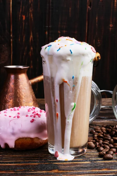 A donut with icing and chocolate powder and a cappuccino glass with high foam and decoration. A can coffee and pouring grains.