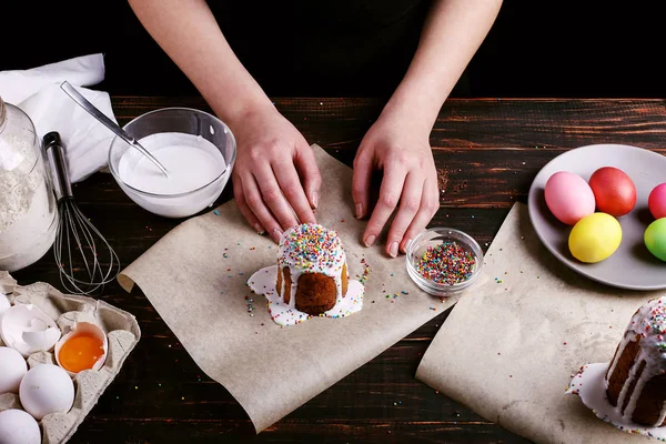 女孩准备复活节烘焙, 用糖霜涂抹蛋糕, 并洒上彩色粉末。在黑暗的背景下, 在厨房的桌子上为假期做准备. — 图库照片