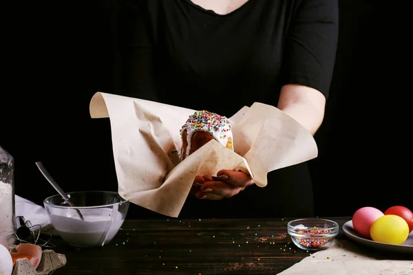 Девушка держит пасхальную выпечку с глазурью и цветным порошком. Подготовка к празднику на кухонном столе, на темном фоне . — стоковое фото