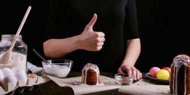 Kız Paskalya pişirme hazırlar, pasta buzlanma ile smear ve renkli toz ile sprinkles. Mutfak masasında karanlık bir arka plan üzerinde tatil için hazırlanıyor.