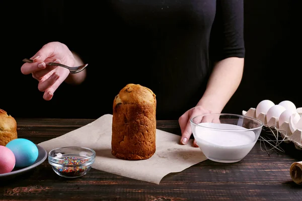 Девушка готовит пасхальную выпечку, намазывает торт глазурью и посыпает цветным порошком. Подготовка к празднику на кухонном столе, на темном фоне . — стоковое фото