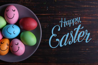 Paskalya yumurtaları plaka üzerinde sevimli yüzü olan. Mutfak masasında karanlık bir arka plan üzerinde tatil için hazırlanıyor. Metin, mutlu Paskalya.