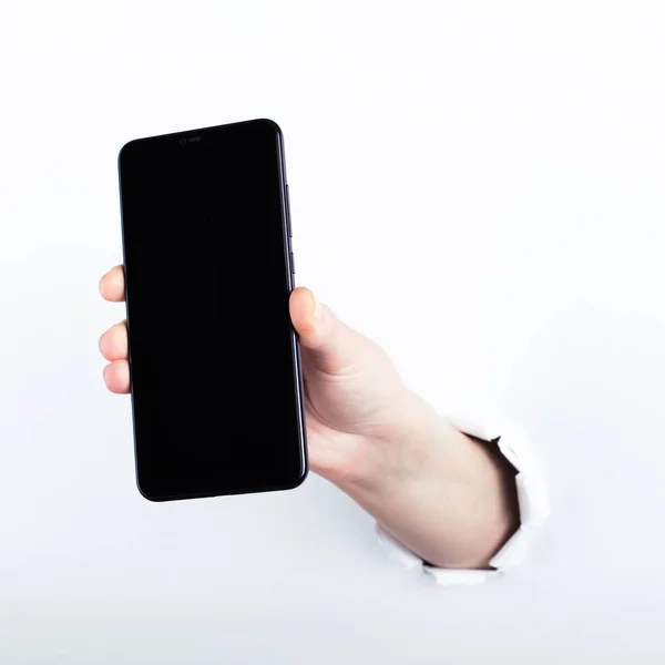 Mano femenina fuera del agujero en el papelero, sosteniendo un teléfono inteligente con una pantalla negra. Aislar sobre fondo blanco . —  Fotos de Stock