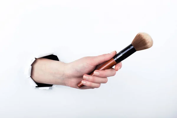 Female hand out of a hole in paper, holding a large powder brush. Isolate on white background. — Stock Photo, Image