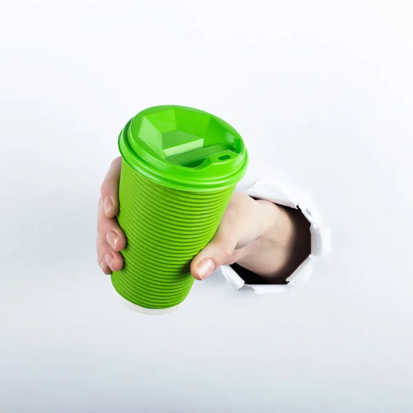 A mão feminina fora do buraco do homem do papel, tem um copo verde de café com ele. Isolar no fundo branco . — Fotografia de Stock